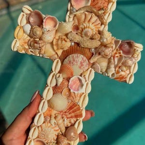 Può includere: Una croce decorativa realizzata con varie conchiglie nei toni crema, pesca e marrone. La croce è adornata con piccole perle e conchiglie di ciprea sui bordi. La croce è tenuta in una mano.