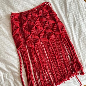 Puede incluir: Una falda de macram&eacute; roja vibrante con un patr&oacute;n geom&eacute;trico de diamantes y largas franjas. La falda est&aacute; hecha a mano con un dise&ntilde;o tejido y presenta una cintura con cord&oacute;n. La franja cuelga de la parte inferior de la falda.
