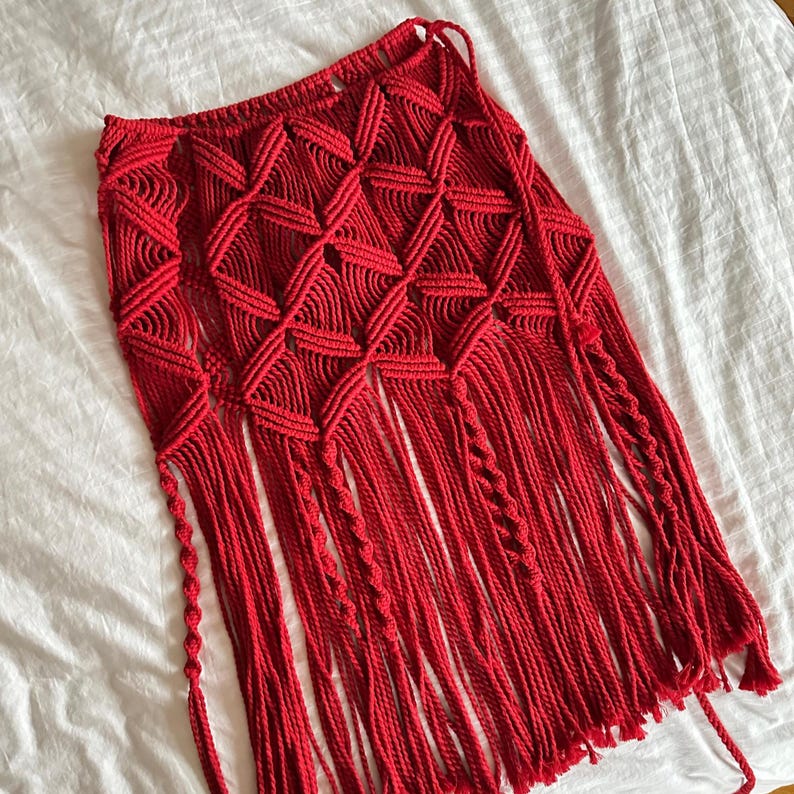Puede incluir: Una falda de macram&eacute; roja vibrante con un patr&oacute;n geom&eacute;trico de diamantes y largas franjas. La falda est&aacute; hecha a mano con un dise&ntilde;o tejido y presenta una cintura con cord&oacute;n. La franja cuelga de la parte inferior de la falda.