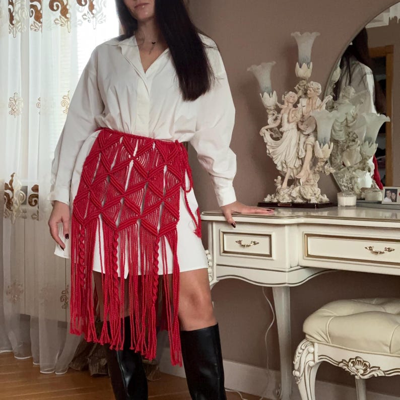 Puede incluir: Una vibrante falda de macram&eacute; roja con un patr&oacute;n geom&eacute;trico de diamantes y flecos. La falda se lleva sobre un vestido blanco y se combina con botas negras. La falda a&ntilde;ade un toque bohemio al conjunto.
