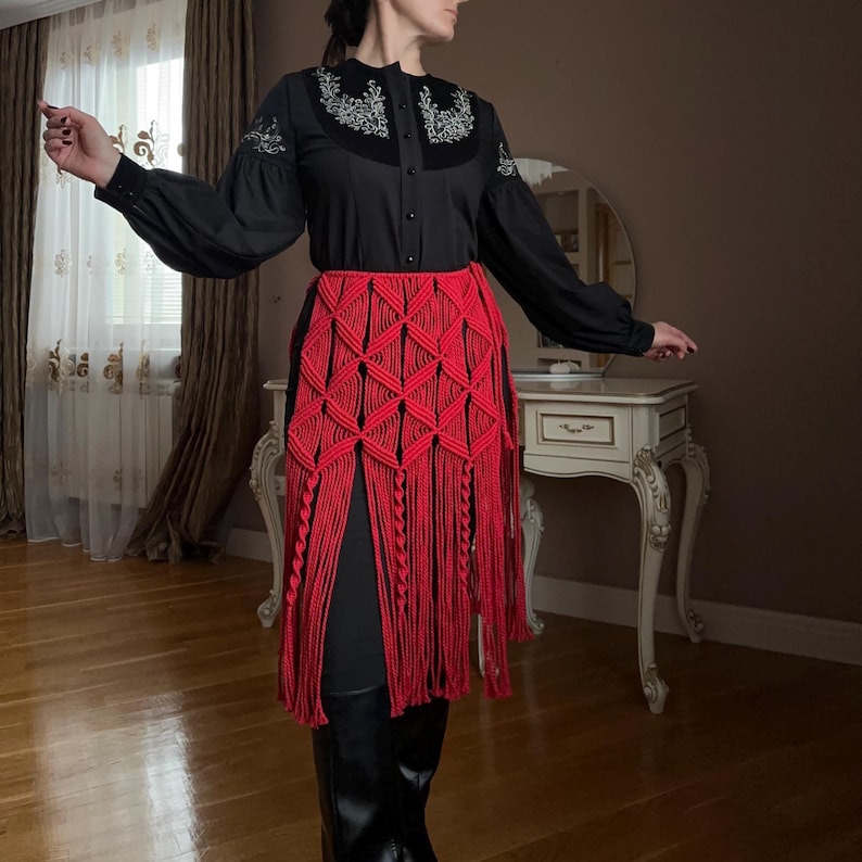 Puede incluir: Una blusa negra con detalles bordados y mangas largas. Una falda de macram&eacute; roja con un patr&oacute;n geom&eacute;trico y flecos se usa sobre leggings negros. El atuendo se combina con botas negras.