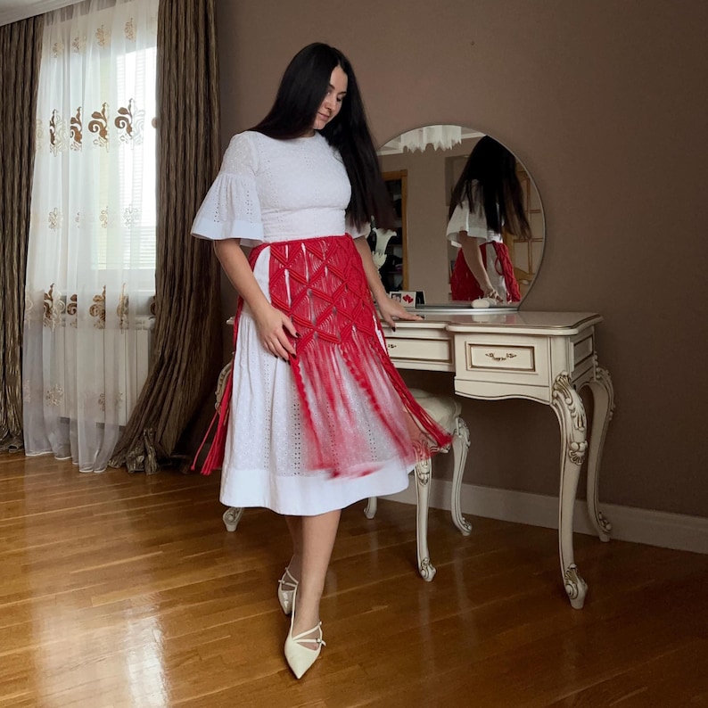 Puede incluir: Conjunto de falda y top blancos con superposici&oacute;n de macram&eacute; rojo. El atuendo se combina con tacones blancos. La habitaci&oacute;n cuenta con un tocador con espejo y suelo de madera.