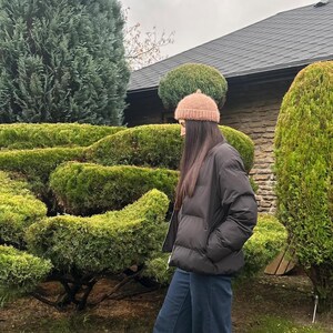 Peut inclure: Une personne portant une veste bouffante noire, un jean bleu et un bonnet en tricot marron clair. L'arrière-plan présente des buissons verts et un bâtiment en pierre avec un toit sombre.