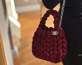 May include: A burgundy-colored handbag with a braided design. The bag has a silver chain strap and a small silver tag. The bag is held by a person wearing a black outfit.
