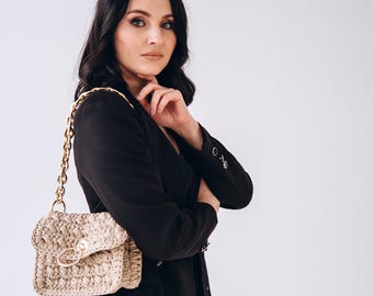 May include: A woman wearing a black blazer with a gold chain strap handbag. The handbag is made of a beige crocheted material.