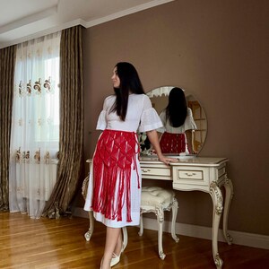 Puede incluir: Una blusa blanca con ojales y mangas cortas y una falda de macram&eacute; roja con flecos. El conjunto se combina con tacones blancos de punta. La habitaci&oacute;n tiene un tocador con espejo redondo y cortina estampada.