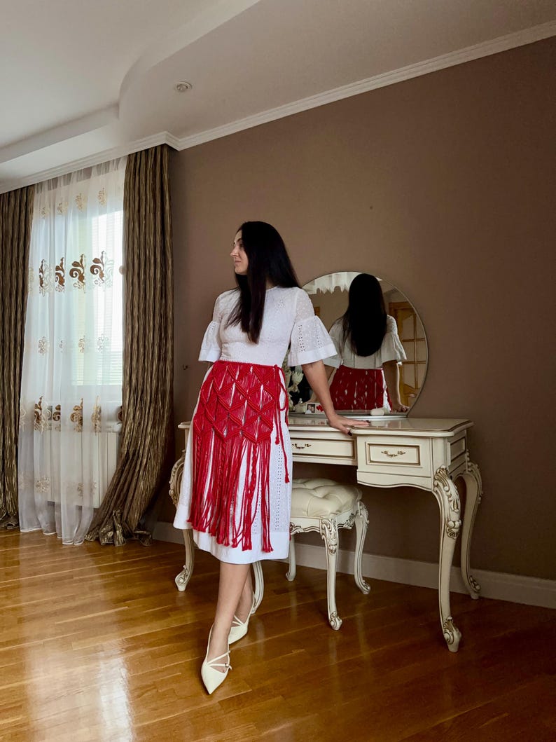 Puede incluir: Una blusa blanca con ojales y mangas cortas y una falda de macram&eacute; roja con flecos. El conjunto se combina con tacones blancos de punta. La habitaci&oacute;n tiene un tocador con espejo redondo y cortina estampada.