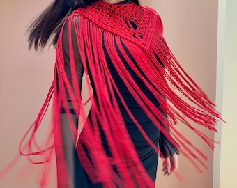 May include: A vibrant red macrame shawl featuring an intricate knotted neckline design and long, flowing fringe that extends over the shoulders and chest, worn over a black long-sleeved garment with sheer mesh sleeves and a solid black bodice.