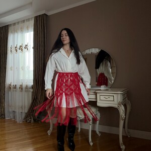 Puede incluir: Una camisa blanca combinada con una falda roja con flecos y botas negras. La falda presenta un dise&ntilde;o tejido con flecos largos. El atuendo se completa con botas negras de punta, creando un contraste audaz.