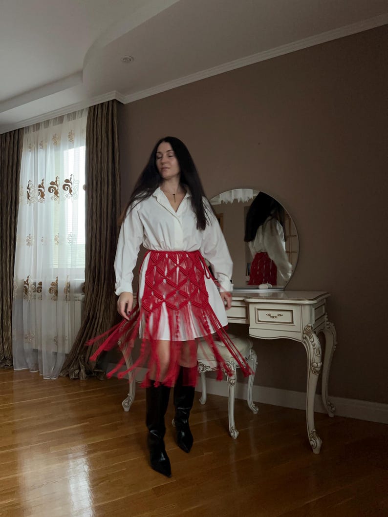 Puede incluir: Una camisa blanca combinada con una falda roja con flecos y botas negras. La falda presenta un dise&ntilde;o tejido con flecos largos. El atuendo se completa con botas negras de punta, creando un contraste audaz.