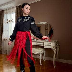 Puede incluir: Una blusa negra con detalles bordados en el cuello y las mangas, combinada con una falda de macram&eacute; roja y leggings negros. El conjunto se completa con botas negras. El estilo general es una mezcla de moda tradicional y moderna.