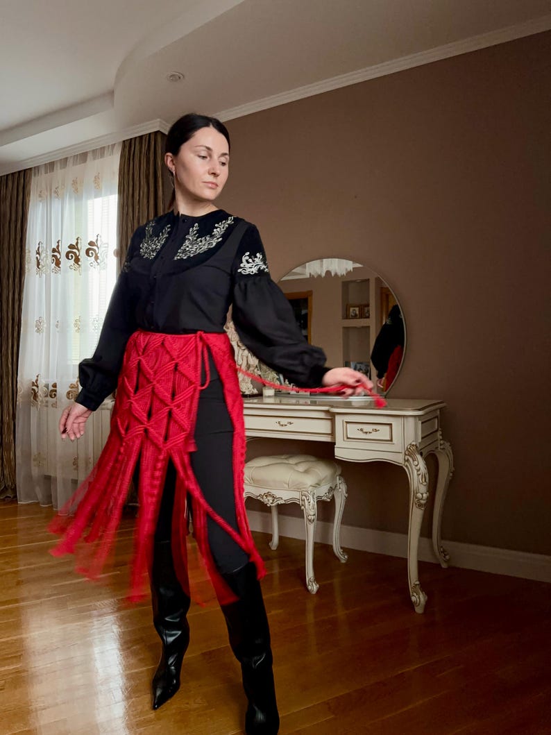 Puede incluir: Una blusa negra con detalles bordados en el cuello y las mangas, combinada con una falda de macram&eacute; roja y leggings negros. El conjunto se completa con botas negras. El estilo general es una mezcla de moda tradicional y moderna.