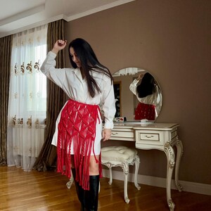 Puede incluir: Una falda de macram&eacute; roja con flecos largos se usa sobre un vestido blanco. La falda presenta un patr&oacute;n geom&eacute;trico y un color vibrante. El atuendo se combina con botas negras hasta la rodilla. El dise&ntilde;o de la falda es el foco principal.