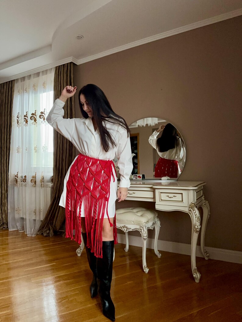 Puede incluir: Una falda de macram&eacute; roja con flecos largos se usa sobre un vestido blanco. La falda presenta un patr&oacute;n geom&eacute;trico y un color vibrante. El atuendo se combina con botas negras hasta la rodilla. El dise&ntilde;o de la falda es el foco principal.