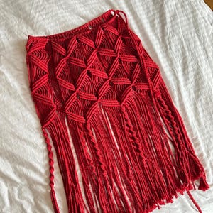 May include: A vibrant red macrame skirt with a geometric diamond pattern and long fringe. The skirt is handmade with knotted rope, showcasing a bohemian style. The skirt is a unique piece of apparel.