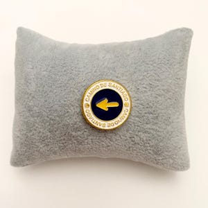 Op de afbeelding: Een ronde metalen speld met een donkerblauw midden en een gele pijl die naar links wijst. De witte rand bevat de goudkleurige tekst Camino de Santiago en twee kleine schelpiconen. De speld wordt gepresenteerd op een klein, rechthoekig grijs kussentje.