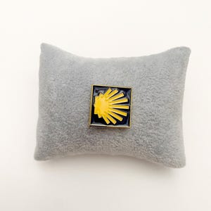 May include: A square enamel pin featuring a yellow scallop shell design set against a dark navy blue background with a thin gold-toned metal border. The pin is displayed on a small, rectangular, light gray plush fabric display pillow.
