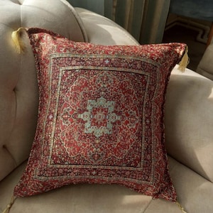 May include: A square decorative pillow with a detailed red and gold pattern, reminiscent of a Persian rug. The pillow has a central medallion design with teal accents and gold tassels at each corner. The pillow is placed on a beige sofa.