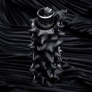 Könnte beinhalten: Ein schwarzes, stacheliges, zylindrisches Objekt mit einem silbernen Oberteil, möglicherweise ein Feuerzeug oder ein ähnlicher Gegenstand. Das Objekt hat eine strukturierte, raue Oberfläche und befindet sich vor einem dunklen, geriffelten Hintergrund.