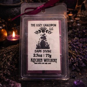 May include: A clear plastic container holding a dark purple wax melt, labeled "Dark Divine" by The Cozy Cauldron LLC. The label includes a cauldron graphic and the text "Kitchen Witchery." The container is tied with a burgundy ribbon, with a dark background.