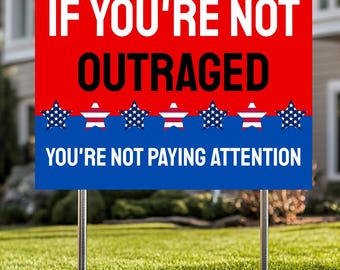 May include: A red, white, and blue yard sign with the text "IF YOU'RE NOT OUTRAGED YOU'RE NOT PAYING ATTENTION." The sign features white stars and stripes, and is mounted on two metal stakes, placed in a grassy yard.