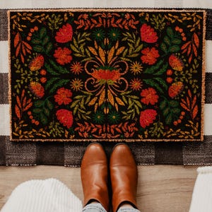 May include: A rectangular doormat with a floral design in red, orange, and green on a black background. The mat is bordered by a black and white checkered pattern. Brown leather boots are visible.