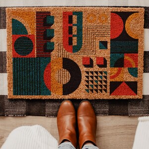 May include: Geometric patterned doormat with a black and white checkered rug underneath. The doormat features a design of colorful shapes including red, teal, orange, and black. Brown leather boots are visible at the bottom of the image, with light blue jeans and white ribbed fabric.