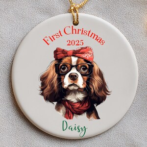 May include: A round, white Christmas ornament with a Cavalier King Charles Spaniel illustration. The dog wears glasses, a red bandana, and a scarf. Text reads "First Christmas 2025" and "Daisy."