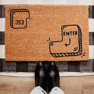 Könnte beinhalten: Eine rechteckige Fußmatte aus natürlicher Kokosfaser. Die Matte zeigt schwarze Strichzeichnungen einer "ESC"-Taste und einer "ENTER"-Taste. Die Matte liegt auf einem schwarz-weiß karierten Teppich, mit den Füßen und schwarzen Stiefeln einer Person sichtbar.