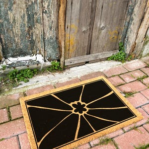 May include: A brown coir doormat with a black geometric design featuring a stylised flower in the centre.