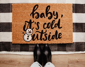 May include: A rectangular doormat with the text "baby, it's cold outside" in black script, accompanied by a small snowman illustration. The mat is brown and sits on a black and white checkered rug, with black boots in the foreground.