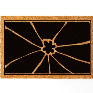 May include: A brown coir doormat with a black floral design. The design features a stylised flower with multiple petals radiating outwards from the centre.