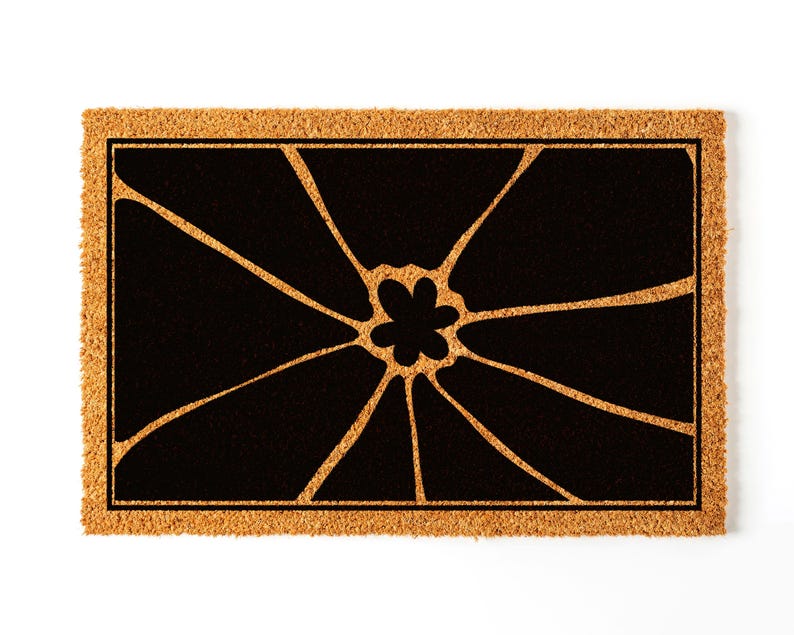May include: A brown coir doormat with a black floral design. The design features a stylised flower with multiple petals radiating outwards from the centre.