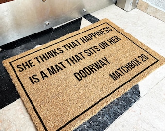 May include: A rectangular coir doormat with a black border and the text "SHE THINKS THAT HAPPINESS IS A MAT THAT SITS ON HER DOORWAY MATCHBOX 20" in black. The mat is placed on a black and white tiled floor.
