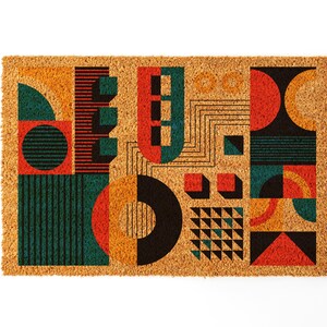 May include: A rectangular doormat with a geometric design. The design features shapes in teal, orange, red, and black. The doormat is made of a natural fiber material.