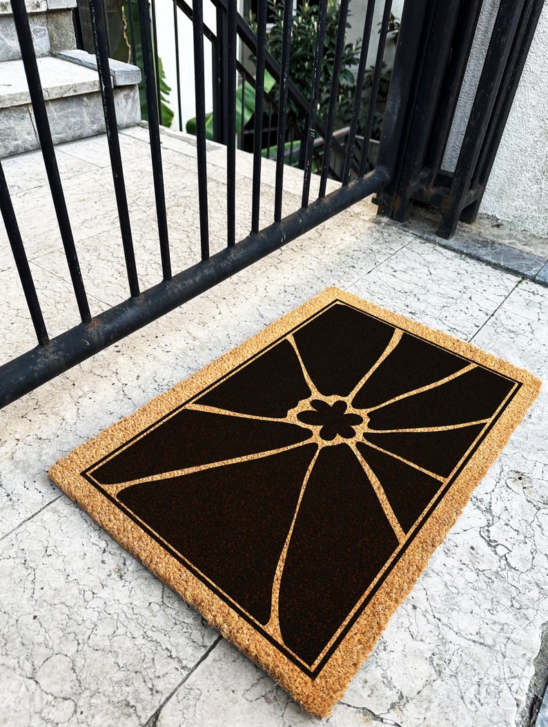 May include: A brown coir doormat with a black geometric design featuring a stylised flower in the centre.