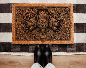 May include: A rectangular doormat with a detailed illustration of a three-headed dog, surrounded by floral and ornamental designs. The artwork is in shades of brown and black, set against a natural coir background. The mat is placed on a black and white checkered rug.