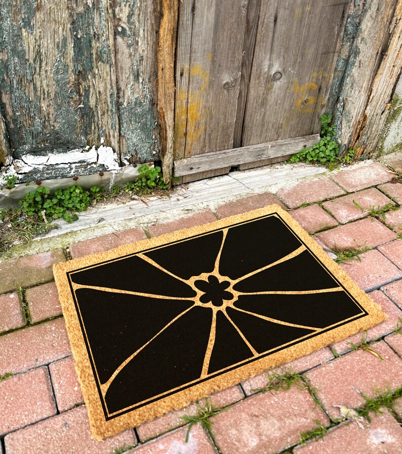 May include: A brown coir doormat with a black geometric design featuring a stylised flower in the centre.