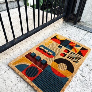 May include: Rectangular doormat with a geometric design. The mat features a tan background with colorful shapes in red, yellow, blue, and black. The design includes circles, squares, and lines. The mat is made of a natural fiber material.