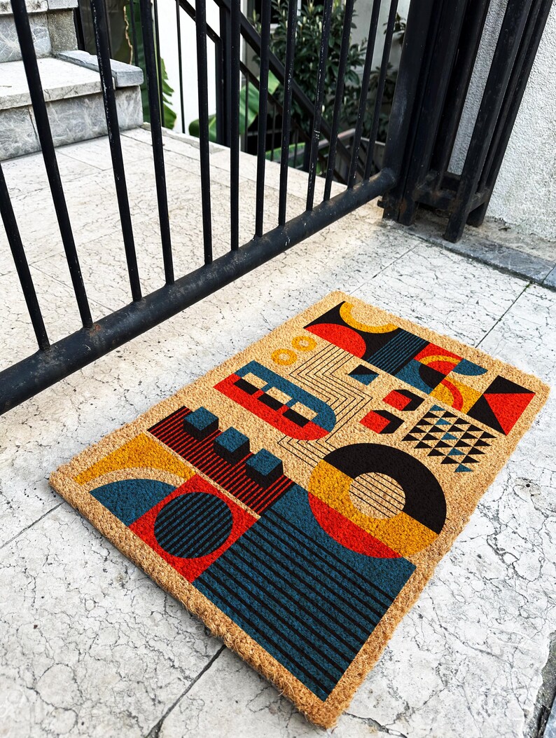 May include: Rectangular doormat with a geometric design. The mat features a tan background with colorful shapes in red, yellow, blue, and black. The design includes circles, squares, and lines. The mat is made of a natural fiber material.