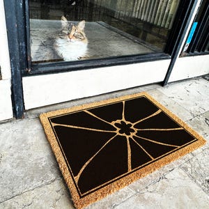 May include: A brown coir doormat with a black geometric design featuring a stylised flower in the centre. The doormat has a natural fibre texture and a rectangular shape.