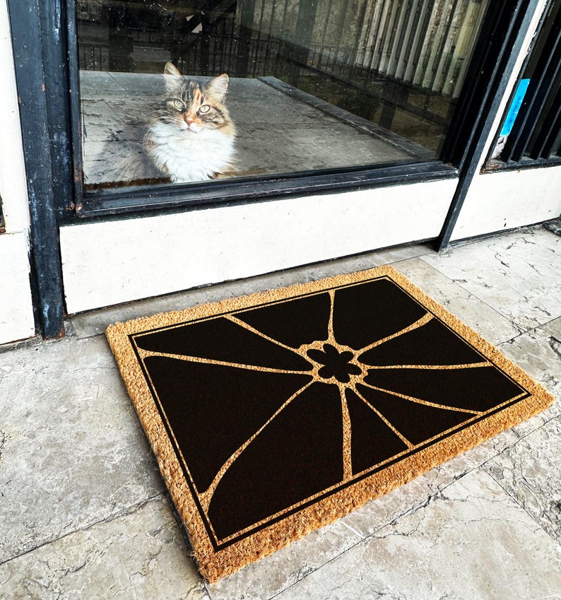 May include: A brown coir doormat with a black geometric design featuring a stylised flower in the centre. The doormat has a natural fibre texture and a rectangular shape.