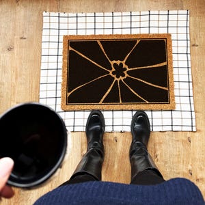 May include: A black and white doormat with a floral design. The doormat is made of coir and has a checkered pattern border. The doormat is on a checkered floor.