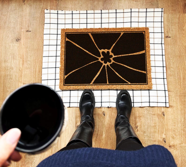 May include: A black and white doormat with a floral design. The doormat is made of coir and has a checkered pattern border. The doormat is on a checkered floor.
