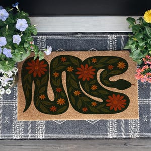 May include: A rectangular doormat with a snake design. The snake is dark green with orange and red floral accents. The mat is on a patterned gray rug, with potted flowers on either side.