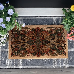 May include: A rectangular coir doormat featuring a symmetrical print of coiled snakes, dark green foliage, and deep orange flowers, layered over a larger dark grey rectangular rug with a white geometric grid pattern and white fringe edges.
