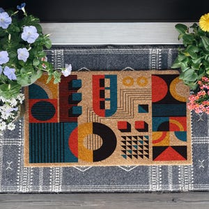 May include: Geometric patterned doormat with a tan background and colorful shapes in red, teal, black, and yellow. The mat is rectangular and sits on a gray patterned rug, flanked by potted flowers.