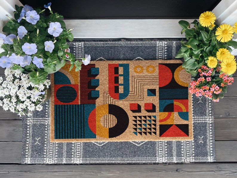 May include: Geometric patterned doormat with a tan background and colorful shapes in red, teal, black, and yellow. The mat is rectangular and sits on a gray patterned rug, flanked by potted flowers.
