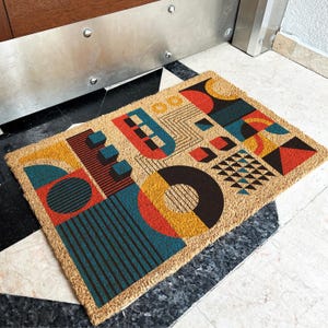 May include: Geometric design doormat with a natural coir fiber base. The mat features a colorful abstract pattern with shapes in red, yellow, teal, and black. The design includes circles, squares, and triangles, creating a modern aesthetic.