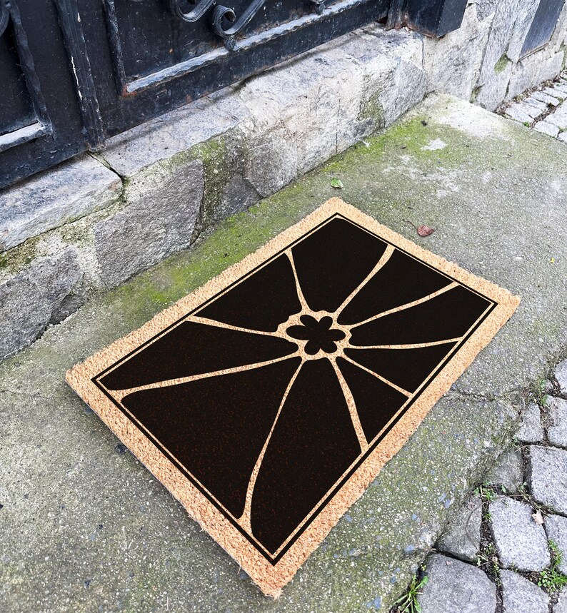 May include: A brown coir doormat with a black floral design. The design features a large flower with eight petals and a smaller flower in the centre.